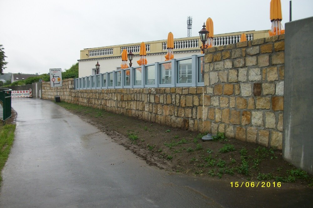 asphaltierter Weg, rechts davon eine Sandsteinmauer mit Fenstereinsätzen, dahinter ein Gebäude