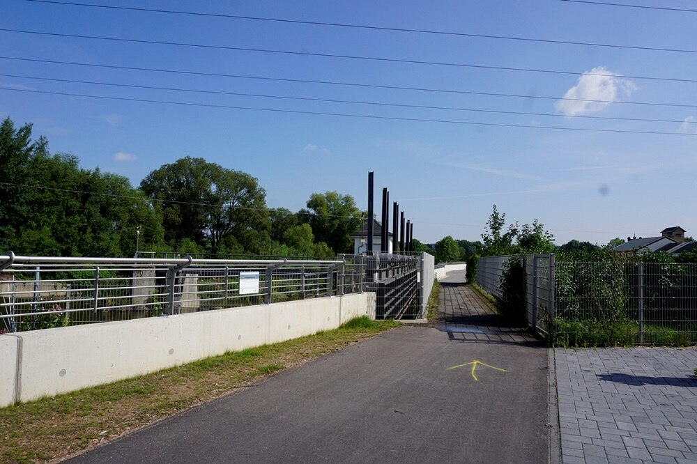 asphaltierter Weg, links niedrige Mauer mit Geländer darauf, blauer Himmel, Bäume