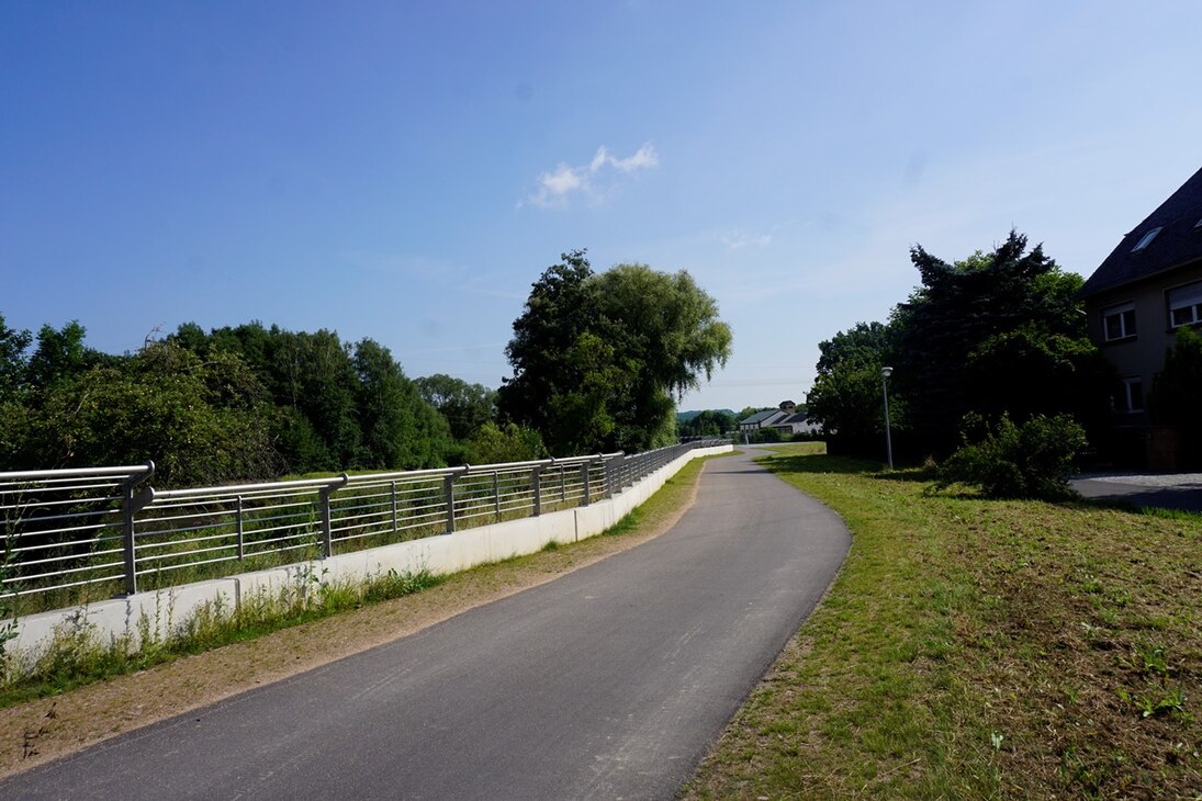 asphaltierter Weg, rechts Wiese, links niedrige Mauer mit Geländer darauf