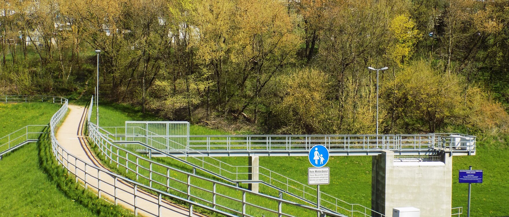 Blick auf eine grüne Wiese durch die ein Weg führt, auf der rechten Seite steht ein Bauwerk mit einem blauen Geländer