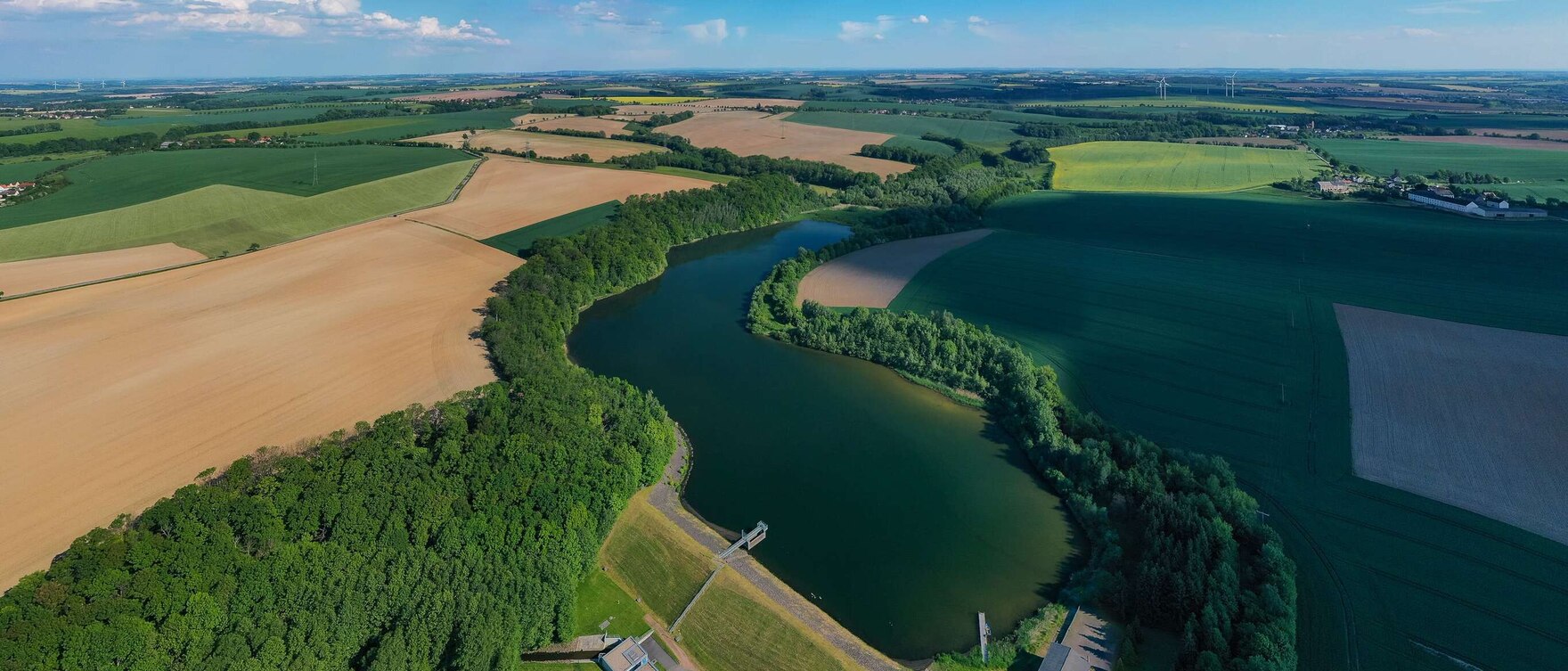 Luftbild einer Talsperre, ringsherum Grün und Felder und im Hintergrund Häuser