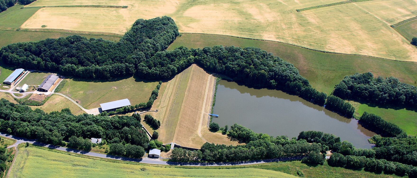 Blick aus der Luft auf einen Stausee mit Damm, umringt von Bäumen und Feldern sowie drei längere Gebäude