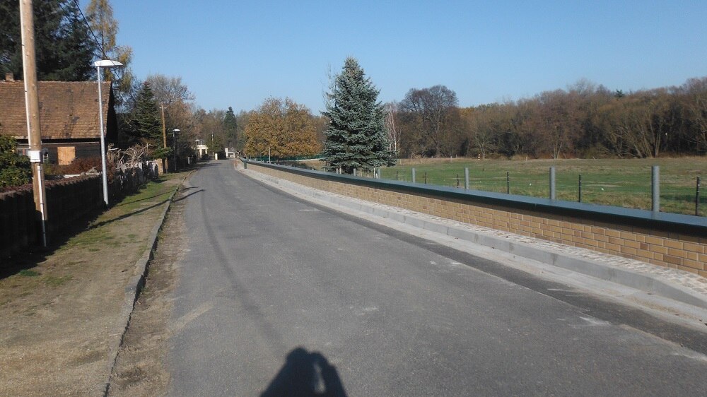 Straße mit niedriger Backsteinmauer auf der rechten Seite, links ein Gehweg, Laternen und Häuser