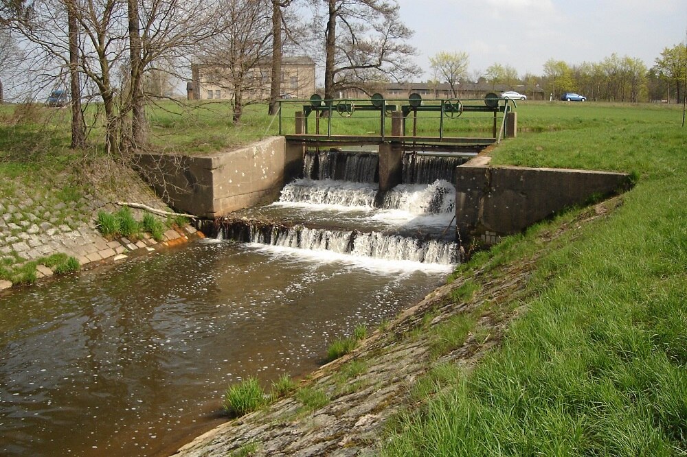 Wasser fließt durch ein altes Wehr, ringsherum grüne Wiese