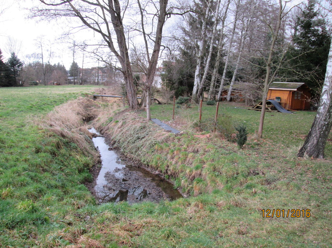 Ein Graben, gefüllt mit ein wenig Wasser führt durch eine Wiese, am rechten Rand stehen Bäume und eine Laube