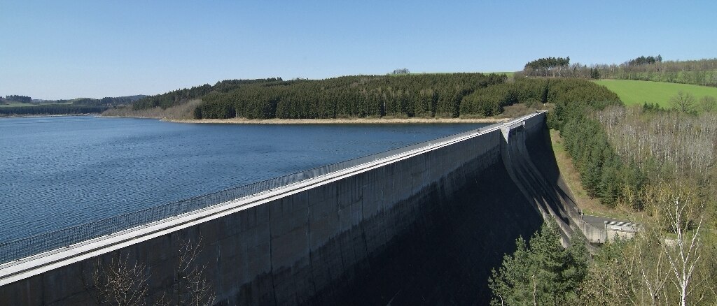 Blick aus der Luft auf die Staumauer mit dem angestauten See, vor der Mauer Bäume