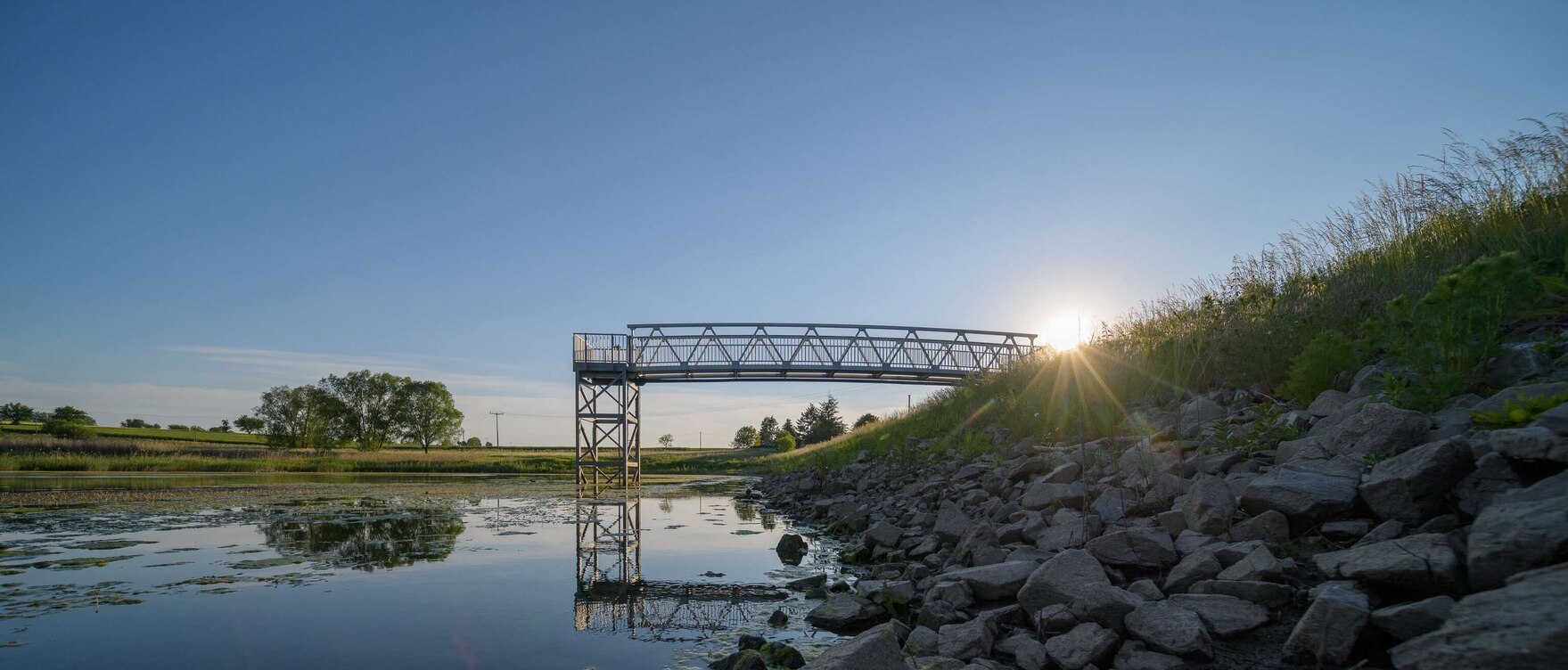 See mit Steinufer und einem Steg aus Metall, der in den See ragt