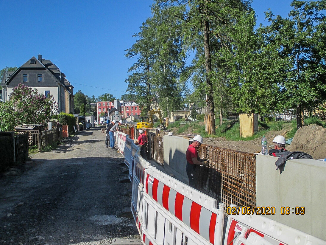 Bauarbeiter mit weißen Helmen stehen hinter einer Absperrung an einer Mauer und arbeiten, daneben eine Straße und im Hintergrund Häuser und Bäume vor blauem Himmel