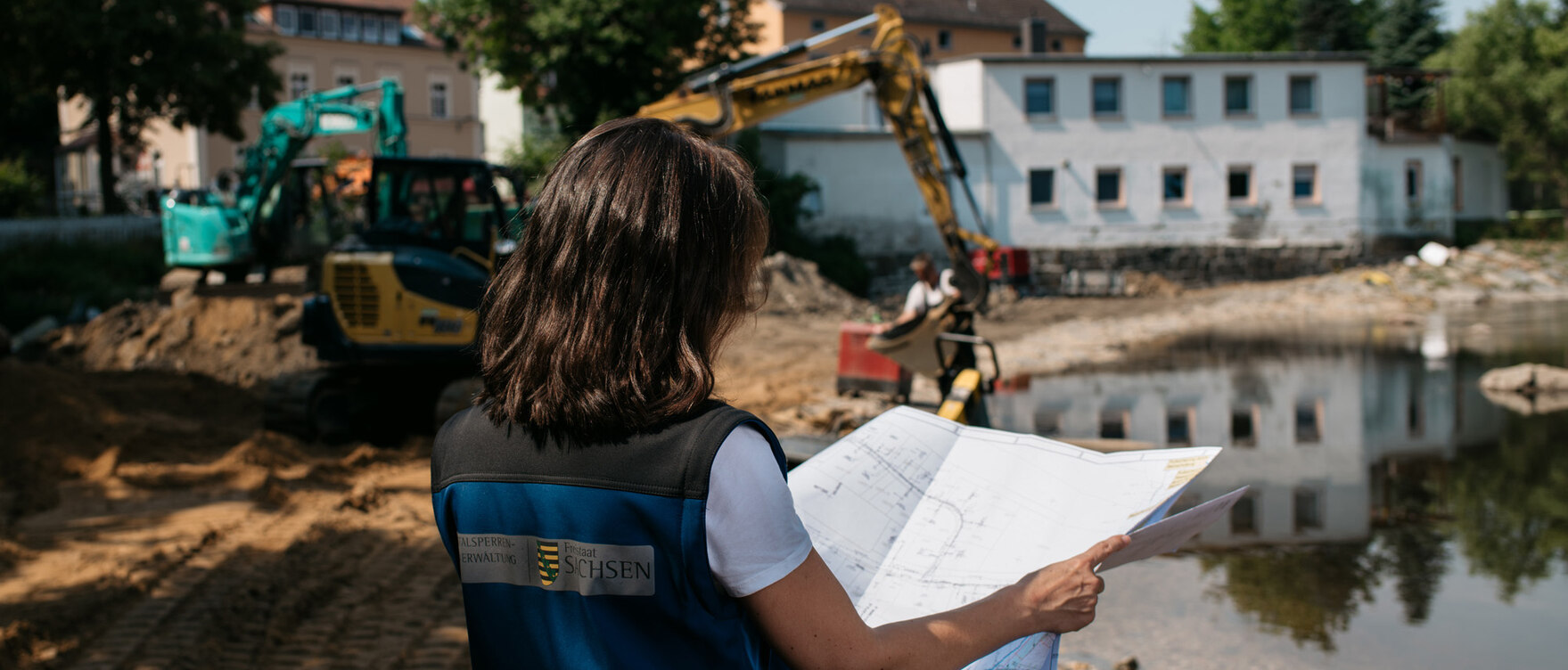 Frau mit einem Plan in der Hand vor einer Baustelle am Fluss